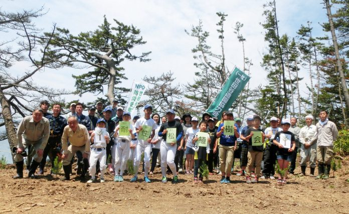 松の木々を背景に、野球のユニフォームを着た子どもたちなどが、「耐性松植樹会」の文字が一文字ずつ書かれた緑色の紙を胸の前に持って前列に並び、後ろに大勢の関係者が並んでいる集合写真