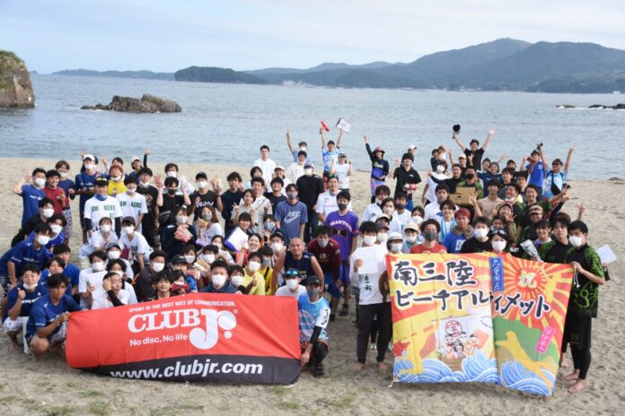 海辺の砂浜で多数の若者たちが集まり、ユニフォームやマスクを着用しながら楽しそうに手を振ったりポーズを取っていて、前方には「CLUB Jr.」と「南三陸ビーチアルティメット」と書かれた2枚の横断幕が掲げられ、背景には穏やかな海と島、遠くの山々が広がっている集合イベントの写真