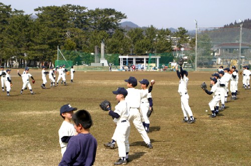 グラウンドで、白いユニフォームを着た複数の少年野球チームの子どもたちが、キャッチボールなどの練習に励んでいる様子を捉えた写真