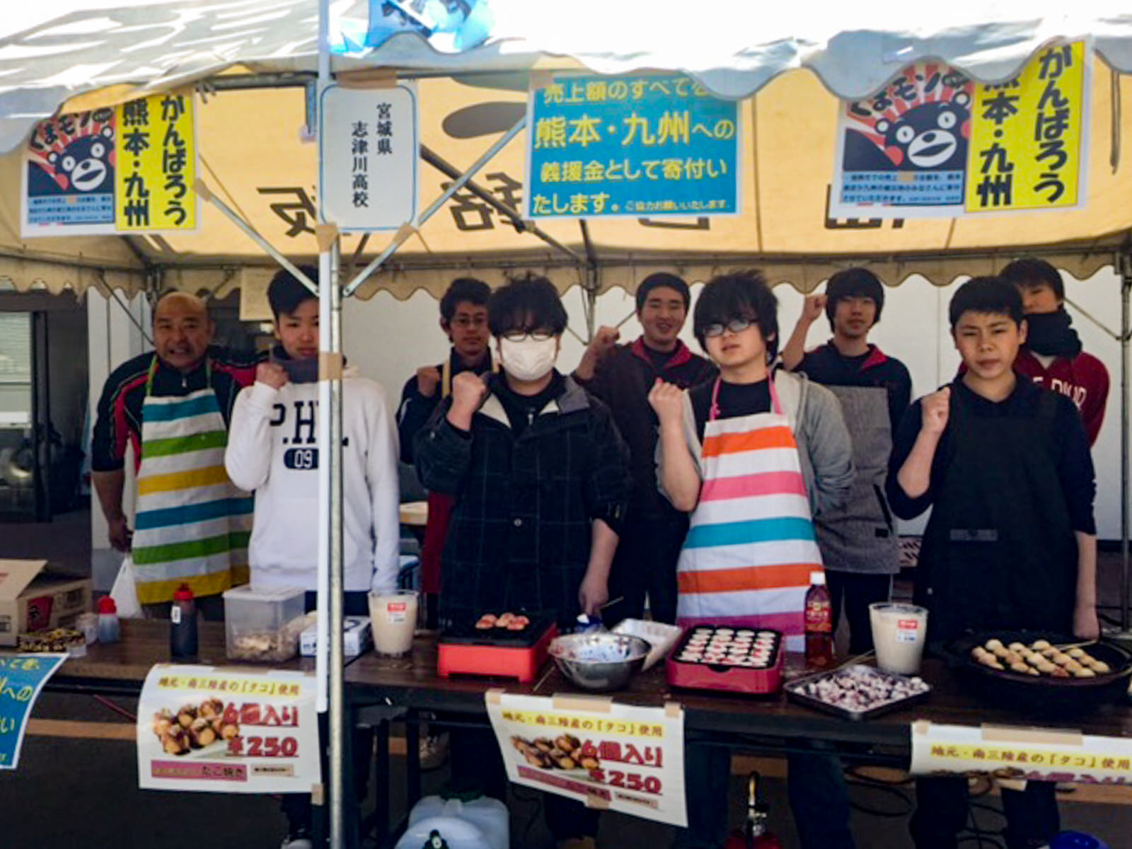 白い屋根のテントの下にタコ焼きのホットプレート3つと、タコ焼きの材料、ソースなどが並んだ長机が置かれており、その後ろに2列に並んだ男子生徒たちがガッツポーズをしている写真