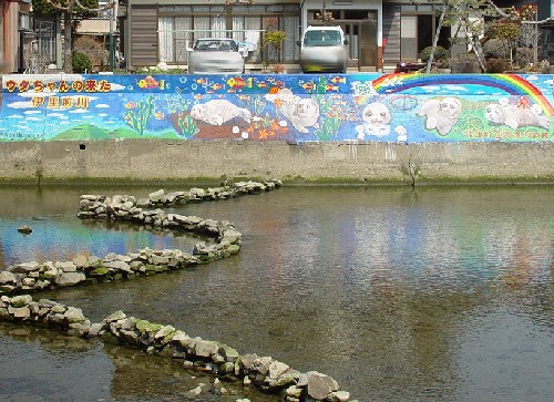 河川敷のコンクリートの壁面に「ウタちゃんの来た伊里前川」と題されたアザラシのウタちゃんや虹が描かれたモザイクアートがあり、その前の浅い川底には蛇行した石組みが配置されている写真