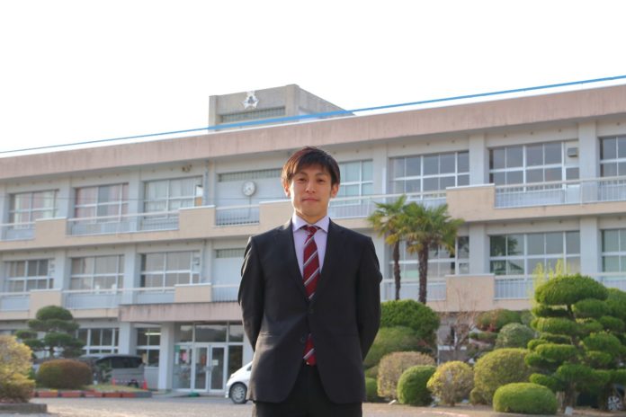 三階建ての校舎と手入れされた植木が見える前で、背広に赤と白のストライプのネクタイを締めた男性がカメラに向かってまっすぐ立っている写真