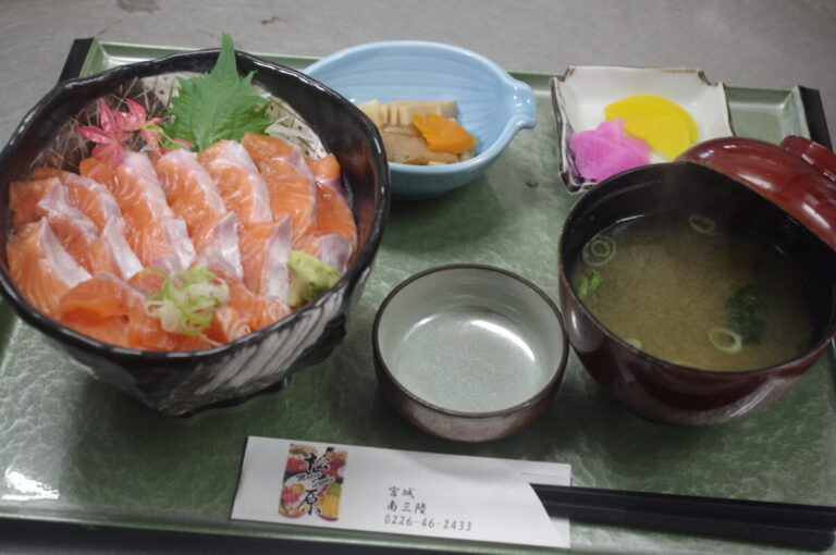 白いご飯の上に厚切りのサケ刺身がきれいに並べられ、ワサビや大葉、ネギとともに盛り付けられ、味噌汁、小鉢、漬物、醤油皿がセットになった、シンプルで上品なサーモン丼の写真