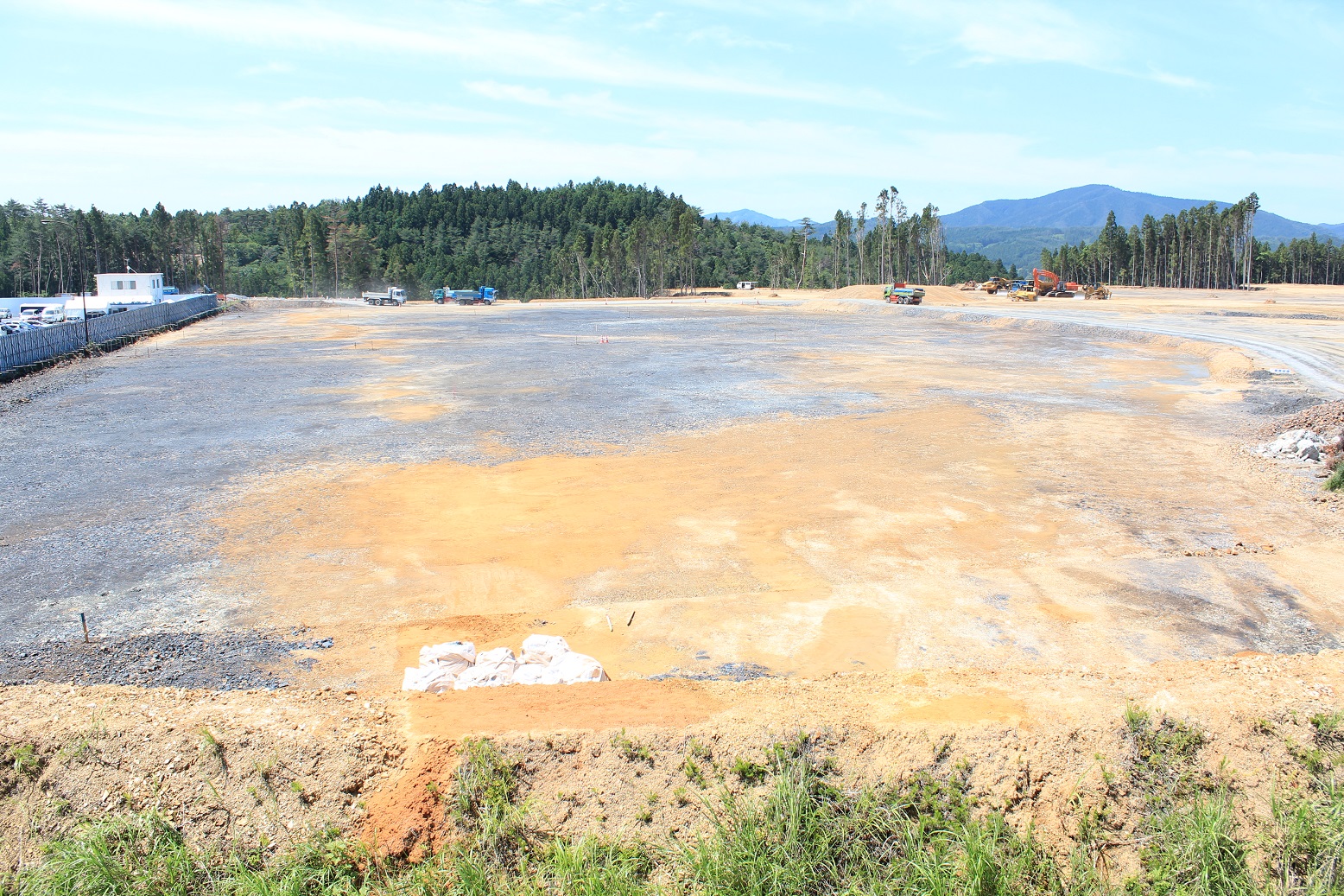 広大な造成地が均されており、手前には白い土嚢が並び、奥には数台のトラックや重機が点在し、背景には緑の森林と山々が広がっている様子の写真