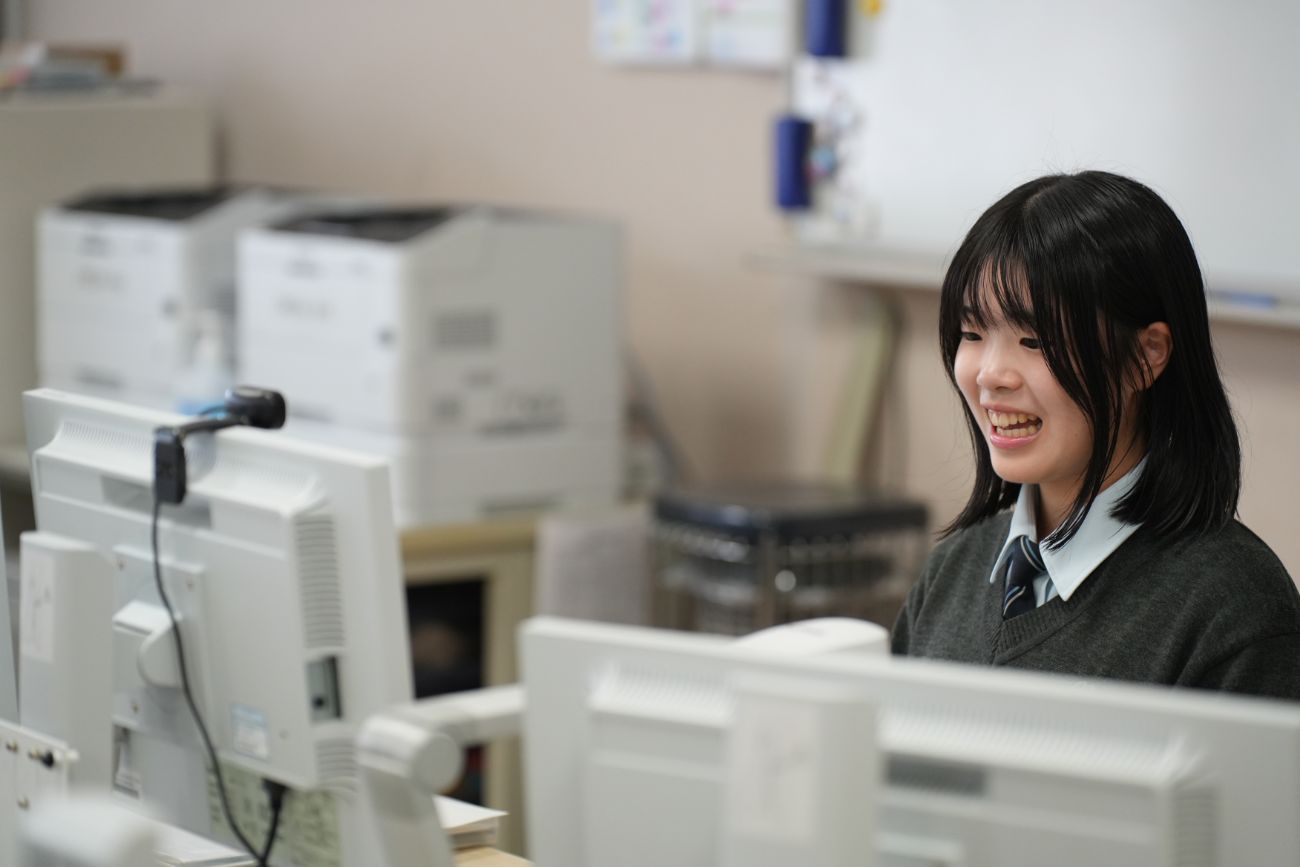 パソコンの前で笑顔で画面を見つめる女子高校生がオンライン授業に参加している様子の写真