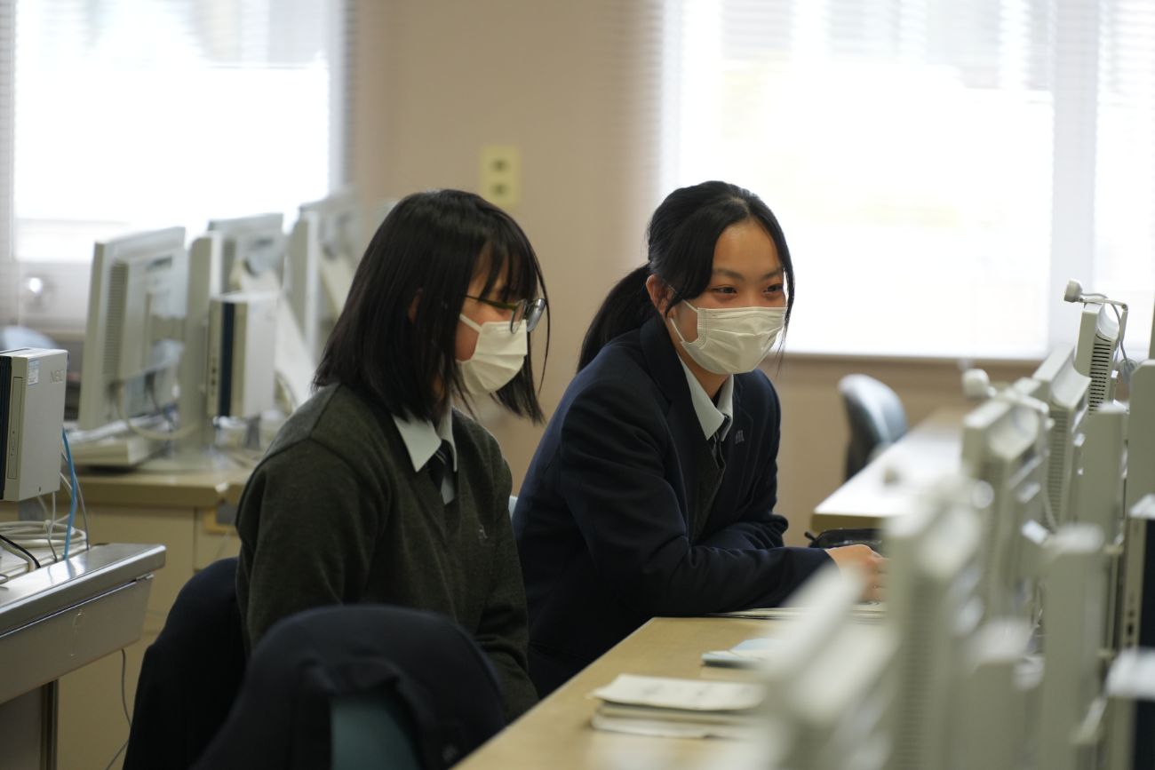 二人の女子学生がマスクを着けてパソコンで作業しながら笑顔で話している様子の写真