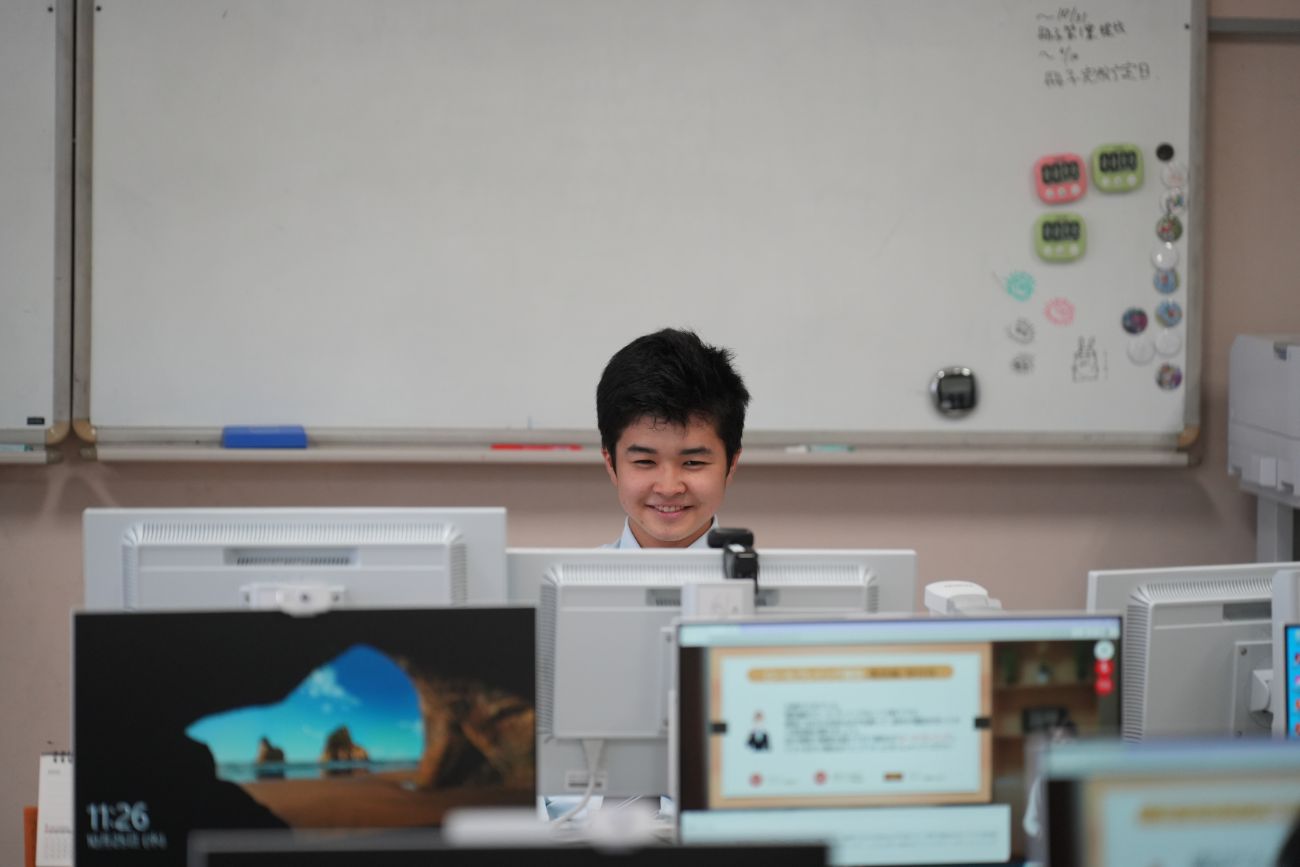 複数のパソコン画面の間からこちらを見て微笑む男子高校生がデジタル学習に取り組んでいる様子の写真