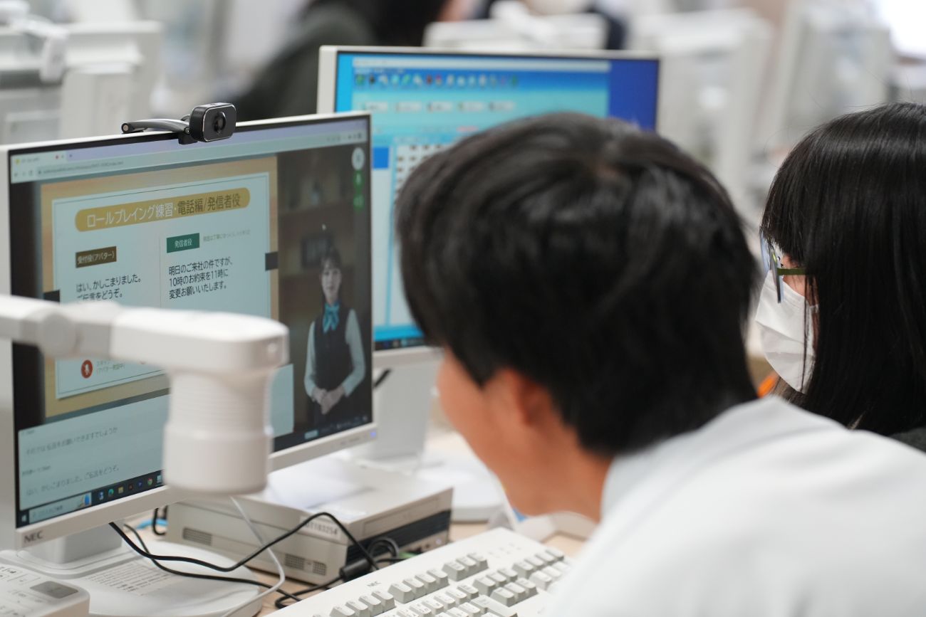 パソコンの画面にビジネスマナーのロールプレイング講習の映像が表示されていて、2人の受講者が画面を見ながらオンライン講義を受けている様子の写真