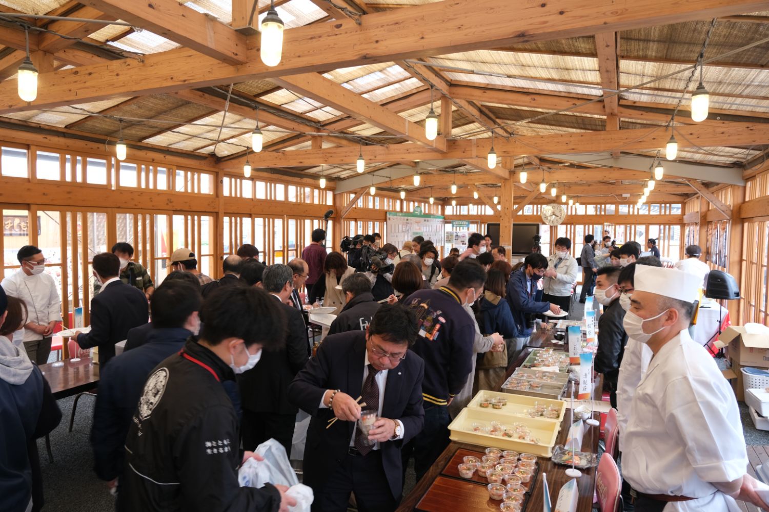 屋内で多くの人が料理の試食や販売コーナーに並び、白衣の料理人が対応している賑やかな様子の写真