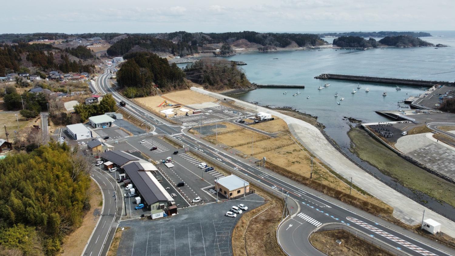 右側にある湾の内側には数十隻の漁船が浮かび、防潮堤と広々とした駐車場、新しい公園、整備された道路が見え、背後には住宅や山が広がる、歌津の海辺と街の海に面した小さな湾とその周辺の集落や山々を一望できる海に面した小さな湾とその周辺の集落や山々を一望できる昼間の風景を高台から定点カメラで撮影した写真