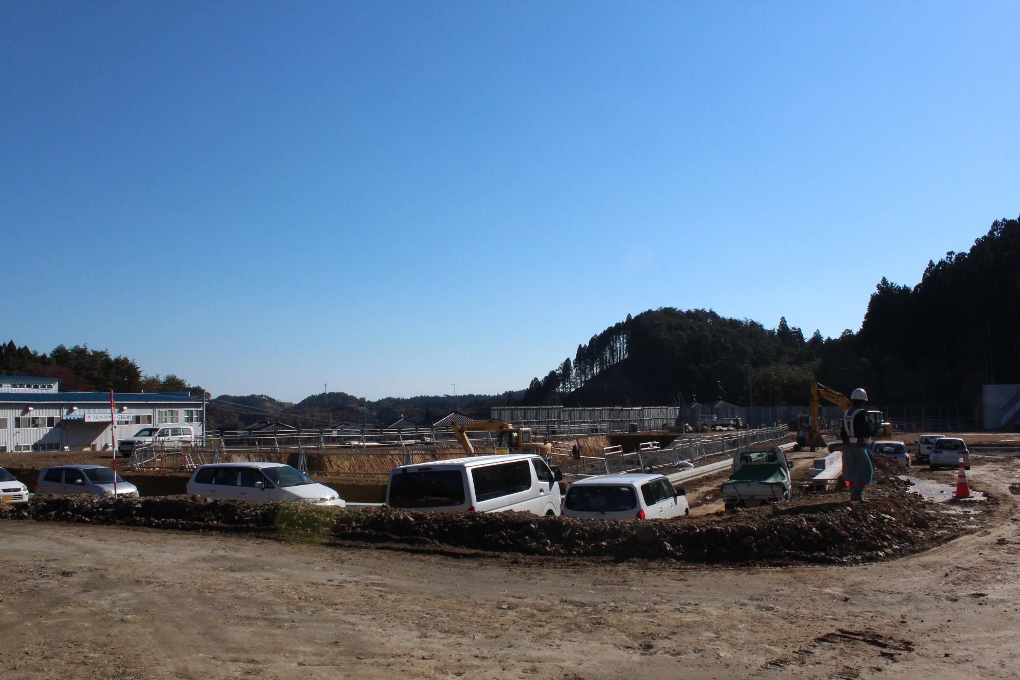 青空の下、建設現場では何台もの白い車両が並び、重機と作業員が基礎工事を進める中、背景には木々に囲まれた小高い山と住宅街が広がる写真