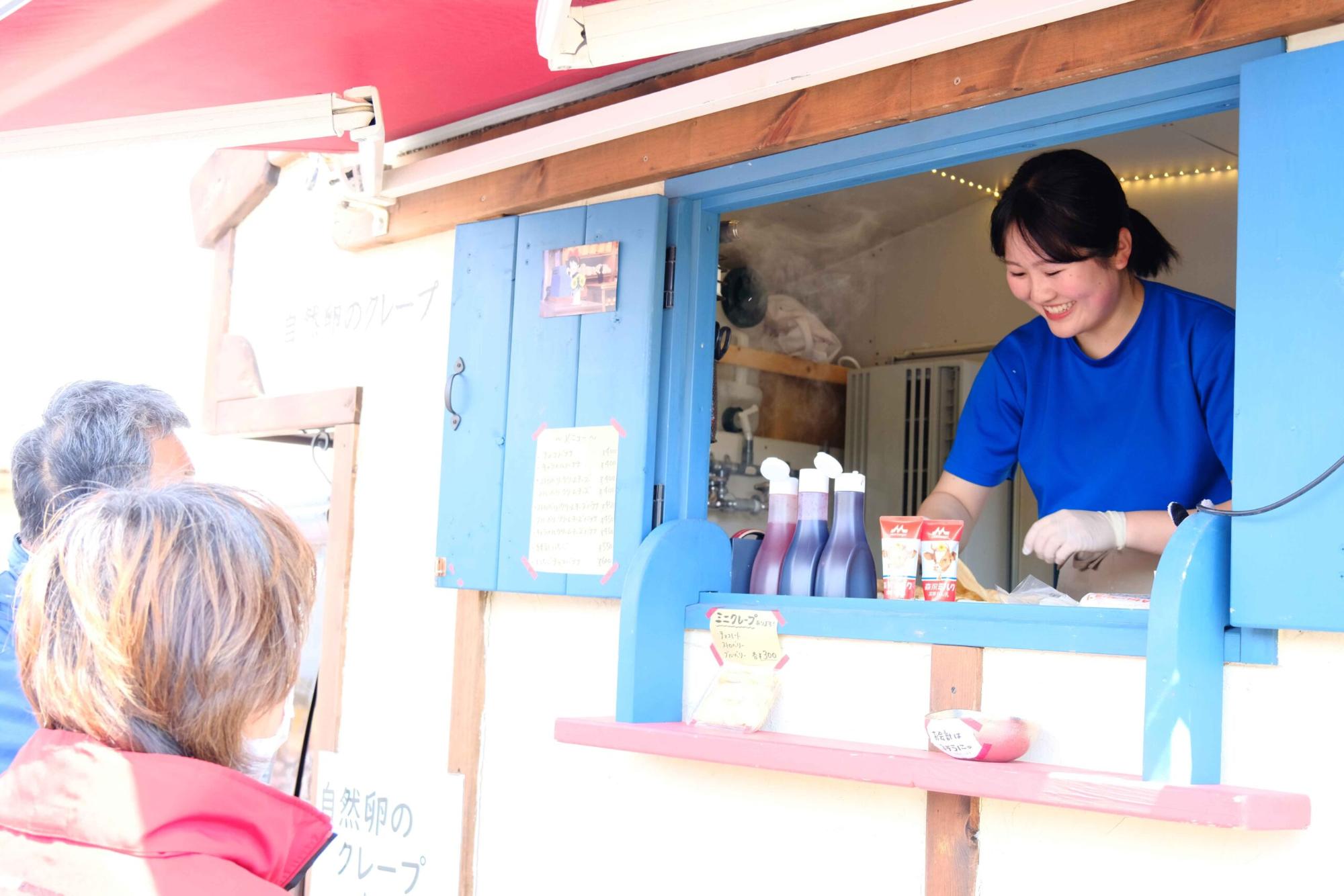 移動販売車の青い小窓からクレープを作る笑顔の女性のが見え、前にはクレープを買い求める来場者が写っている写真