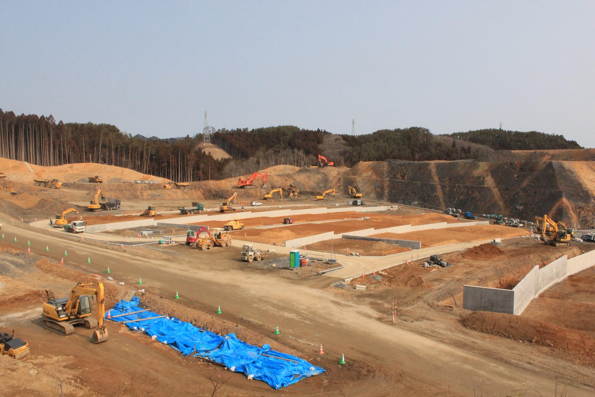 高台部分の土地が均され、写真中央部には、青いシートで覆われた資材置き場、仮設フェンスや資材ヤードがあり、造成地に沢山のショベルカーなどの重機が配置されている写真