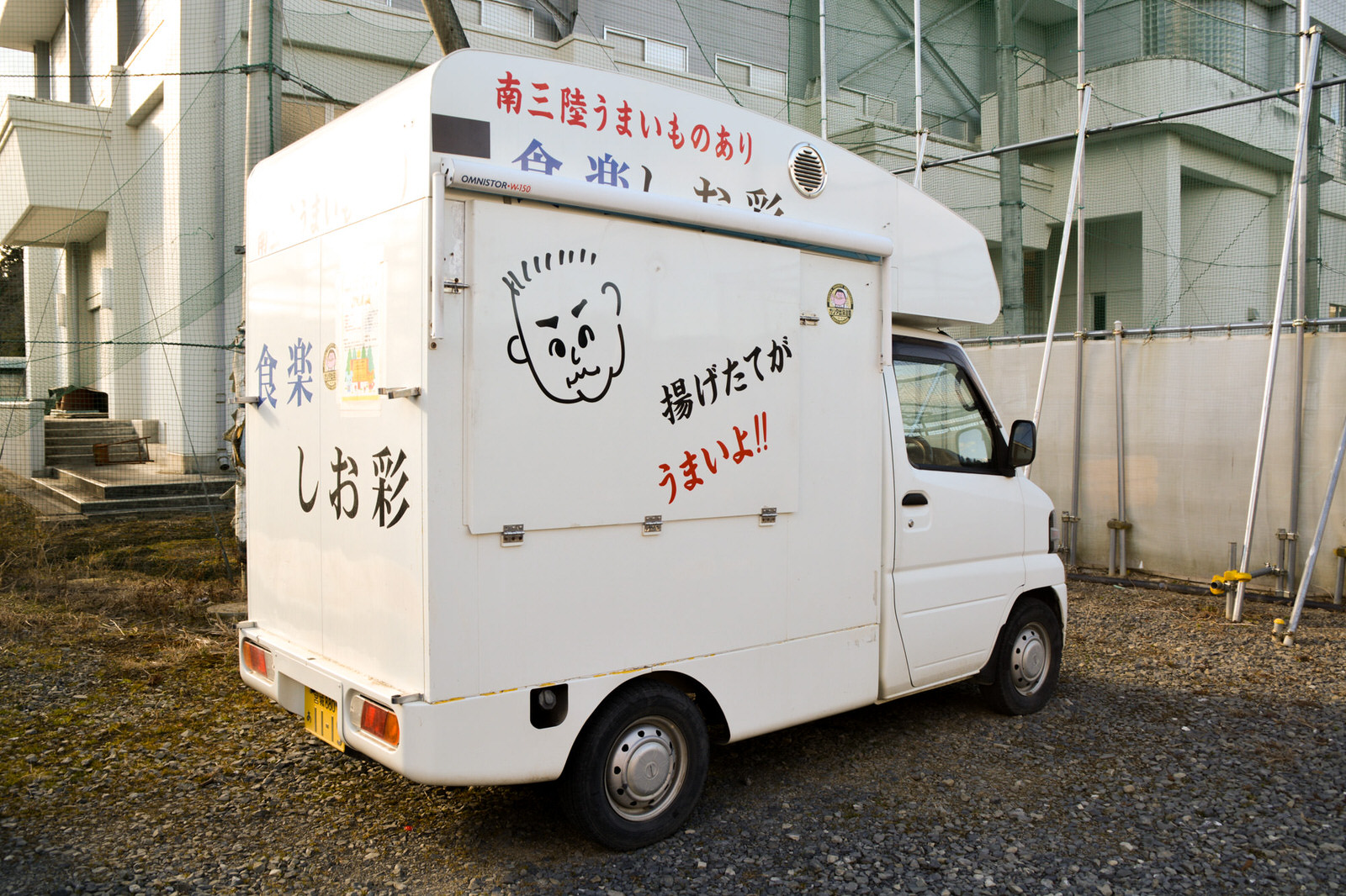 側面に男性の似顔絵と「揚げたてがうまいよ！！」の文字が書かれ、後ろに大きく「しお彩」と書かれた白い移動販売車が砂利の駐車場に停まっている写真