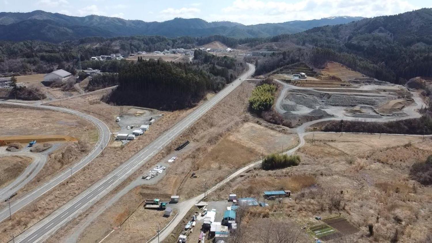 中央を縦に国道がまっすぐ走り、その両側には造成中の土地や緩やかな山の斜面が広がっていて、左には曲がりくねった市道と仮設のような建物群、右の山裾には削られた土の段差が見え、遠くには森林と復興中の集落が点在している戸倉地区内陸部の昼間の風景を高台から定点カメラで撮影した写真