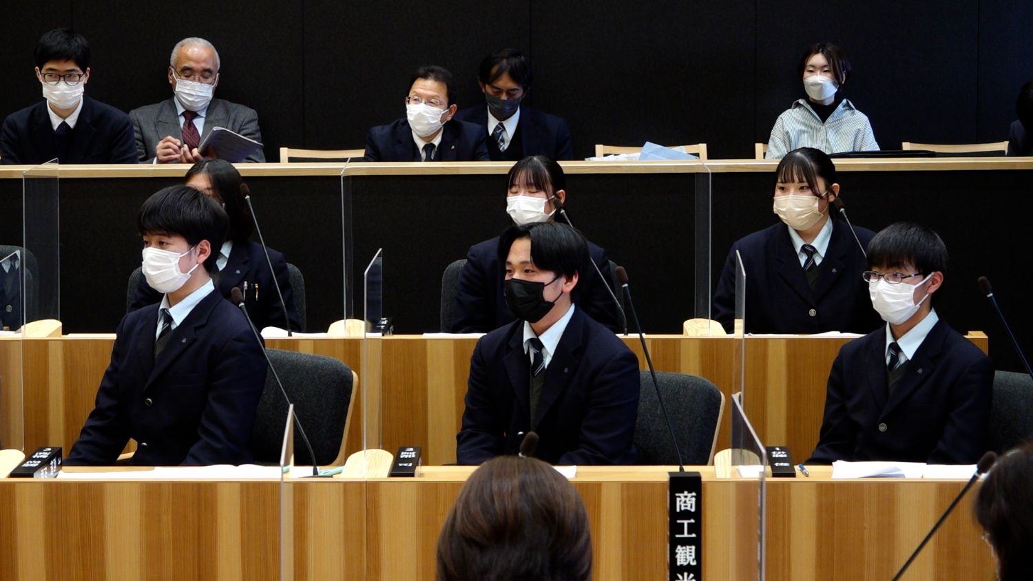 マイクが並ぶ議場の中で、前方に制服姿の中高生たちがマスクを着けて緊張した面持ちで座り、後方の傍聴席にはスーツ姿の大人や私服の人々が資料を見たり見守ったりしている写真