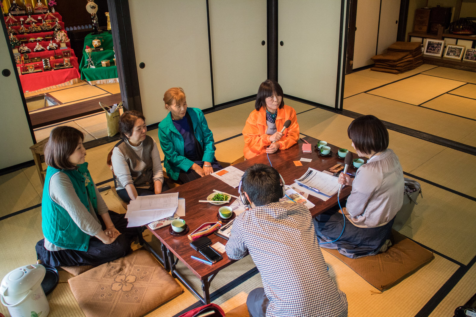 雛飾りが飾られた部屋の隣の和室で、5人の女性とヘッドホンをした男性1人がテーブルを囲み、2人の女性がマイクを持ってインタビューを行っている様子の写真