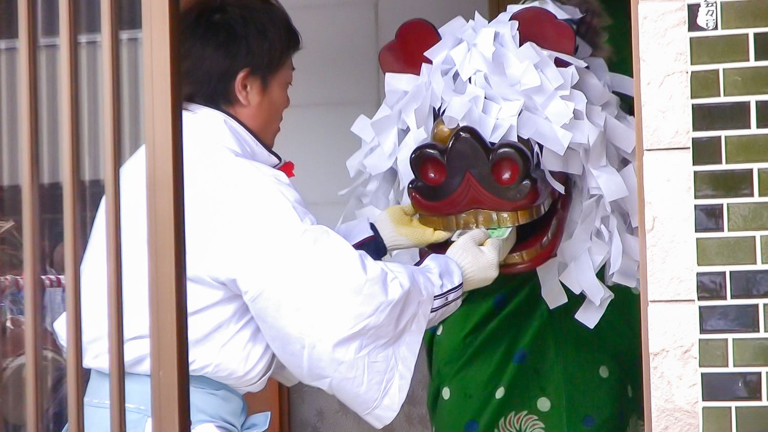 玄関先で白装束の男性が、白い紙飾りを頭につけた獅子舞の口にパック入りの豆腐を入れている様子の写真