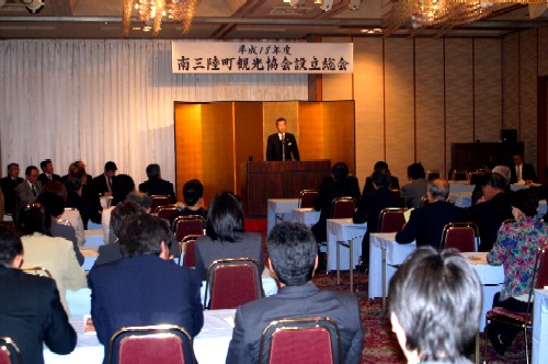 広い宴会場のような場所で、スーツ姿の演説者が金屏風の前にある演台でスピーチをしており、多くの参加者が席について熱心に話を聞いている、南三陸町観光協会設立総会の様子の写真