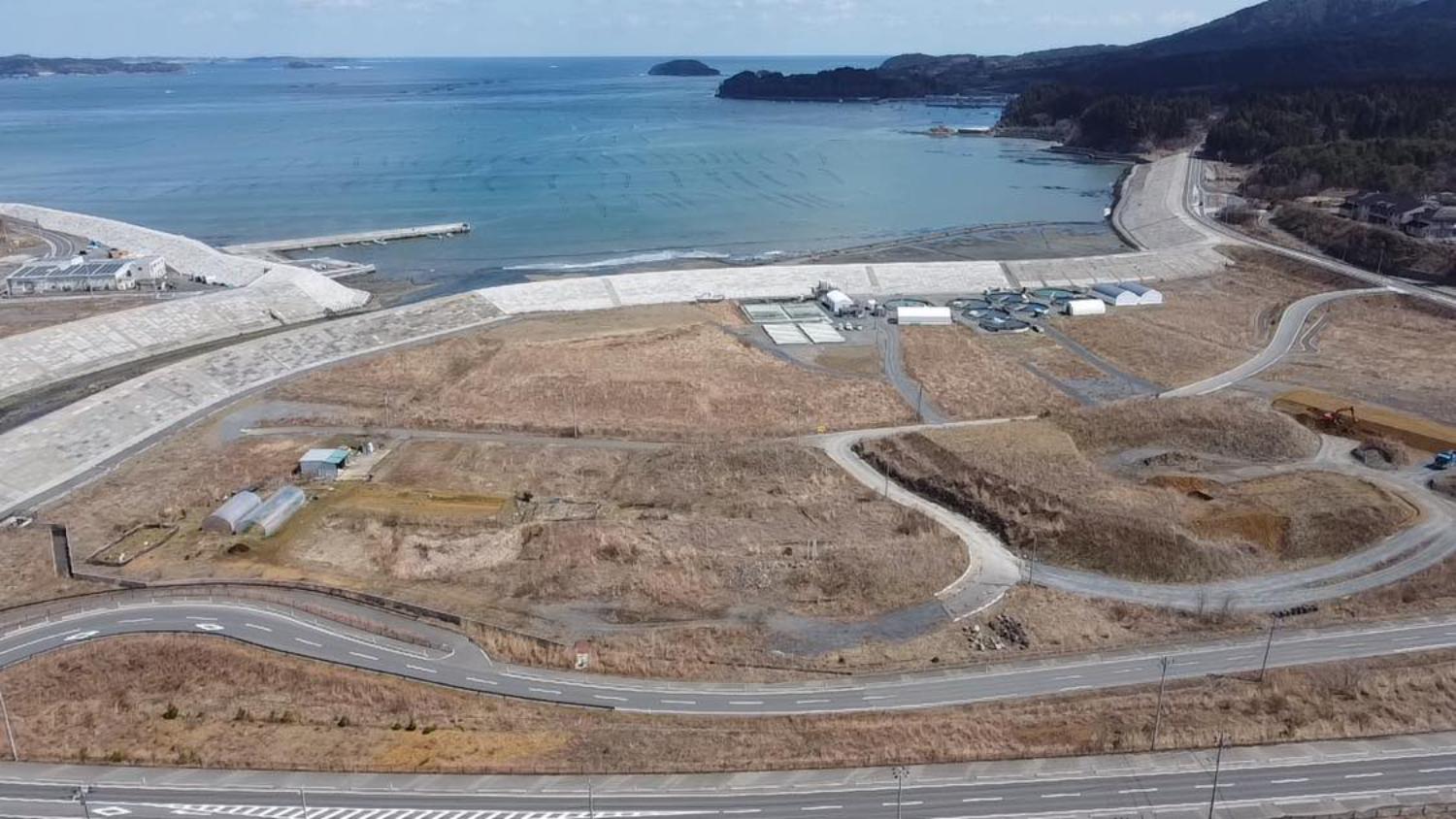 内陸に向かってまっすぐ伸びる国道と、その左右に広がる造成された土地や曲がった支道、奥には山間の集落や造成中の斜面があり、再開発の進む様子が見て取れる内陸部の昼間の風景を高台から定点カメラで撮影した写真