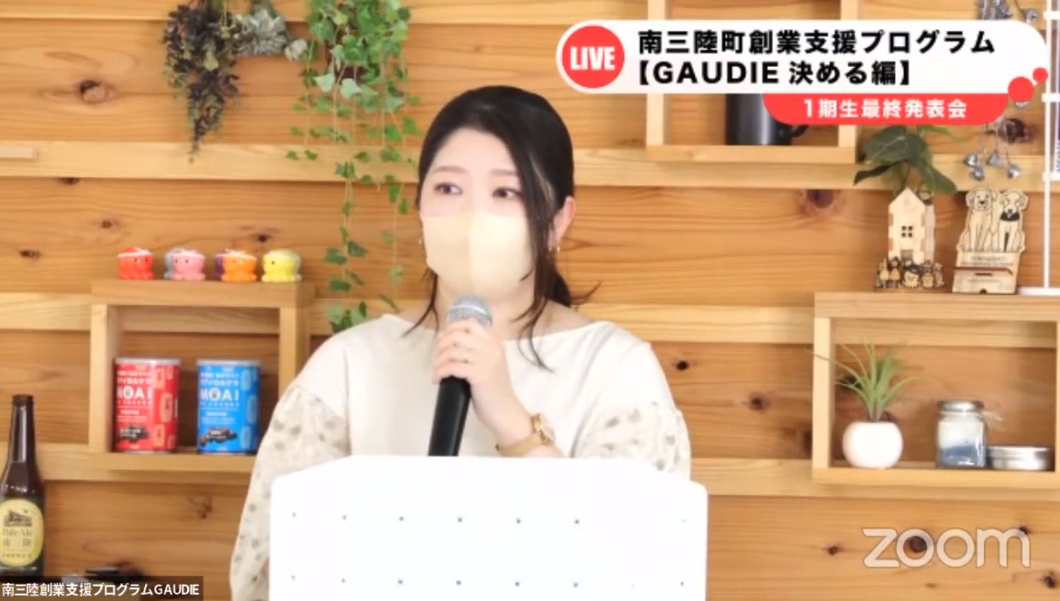 生成り色の服を着た女性がマスクを着けてマイクを持ち、ビール瓶や植物が飾られた棚の前でGAUDIEの発表会でスピーチしている様子の写真
