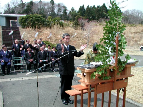 背後にスーツ姿の参列者が座る屋外の式典で、関係者の男性が玉串が供えられた祭壇の前で拍手をしようとしている神割崎キャンプ場開きの様子の写真