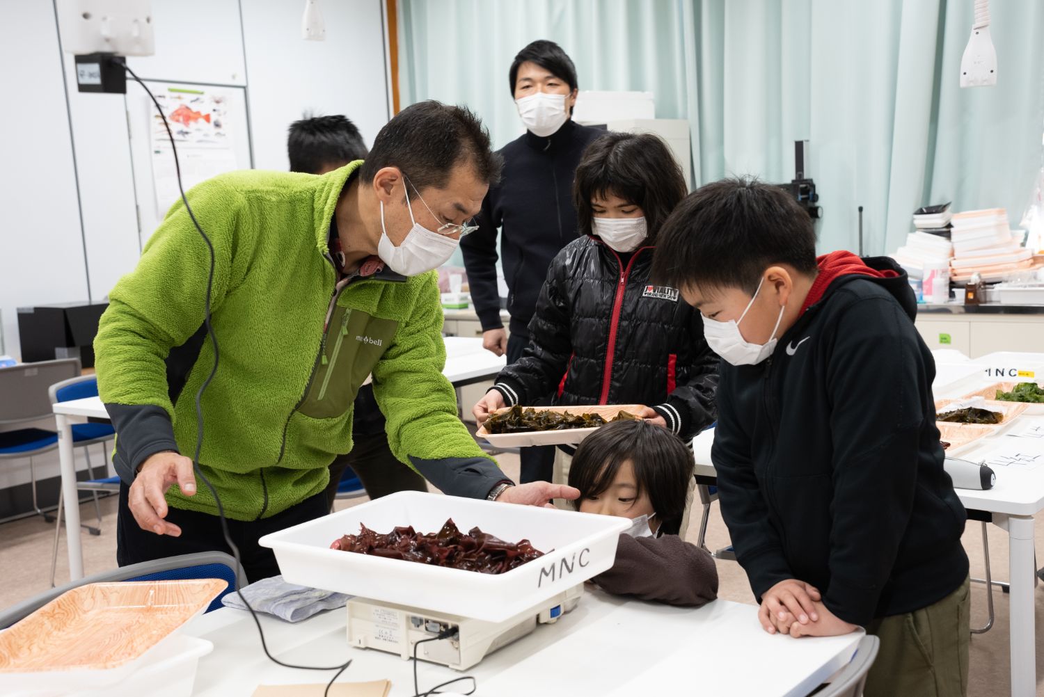 関係者の男性がトレーの海藻を量りにのせて測定し、子どもたちが興味深そうに見つめている様子の写真