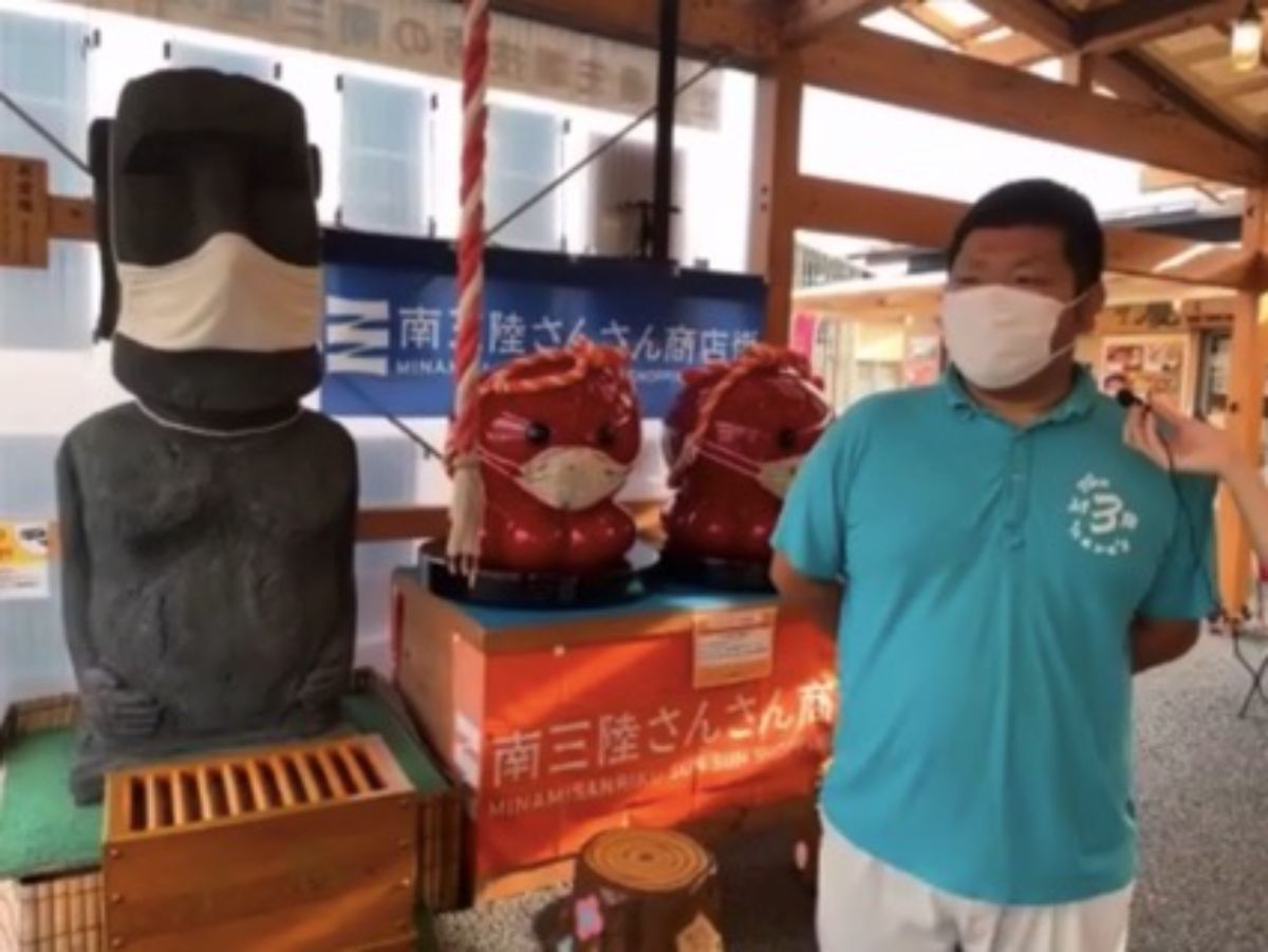 商店街の室内にあるモアイ像と2体の赤いオクトパス君がそれぞれ白いマスクを着用して並べられており、その横で青いポロシャツを着た男性がインタビューを受けている様子のスクリーンショット画像