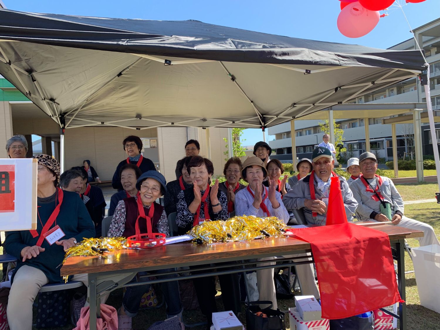 テントの下にテーブルには赤い布や装飾品が置かれ、首に赤いハチマキを巻いたメンバー達が応援する様子の写真