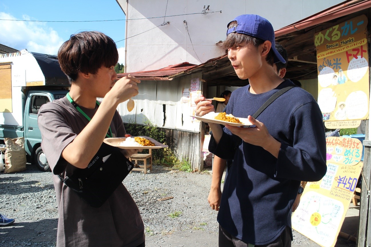 屋外で男性2人が並んで立ち、それぞれの皿に盛られたカレーライスをスプーンで食べている様子が写っている写真