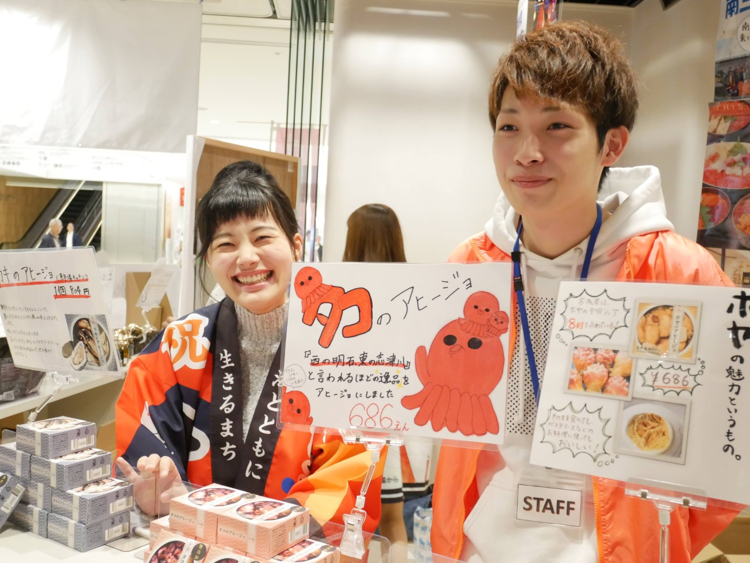 「海とともに生きるまち」と書かれたカラフルな法被を着た女性スタッフが満面の笑みで接客しており、隣に立つ男性スタッフとともに「タコのアヒージョ」などの手描きポップや商品を並べた明るい物産展の売り場の様子を写した写真