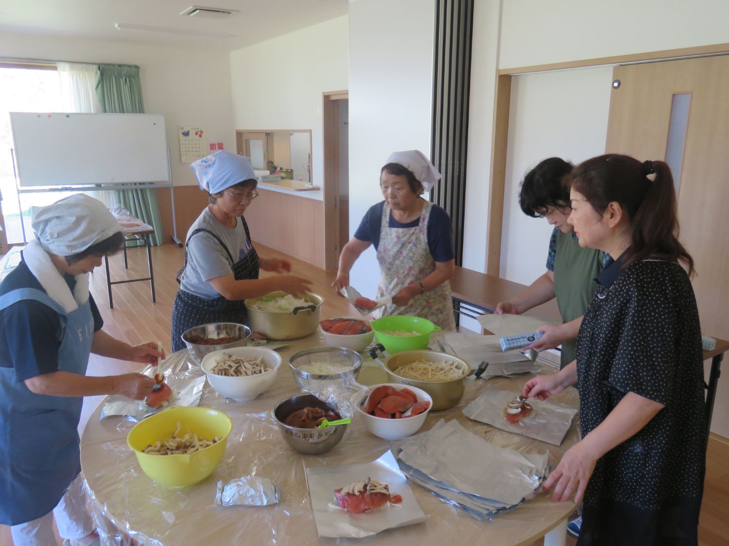 明るい室内で、エプロンや三角巾を着けた女性たちが円卓を囲み、アルミホイルの上に野菜やソーセージなどの具材を丁寧に並べて料理の仕込みをしている様子が写された写真