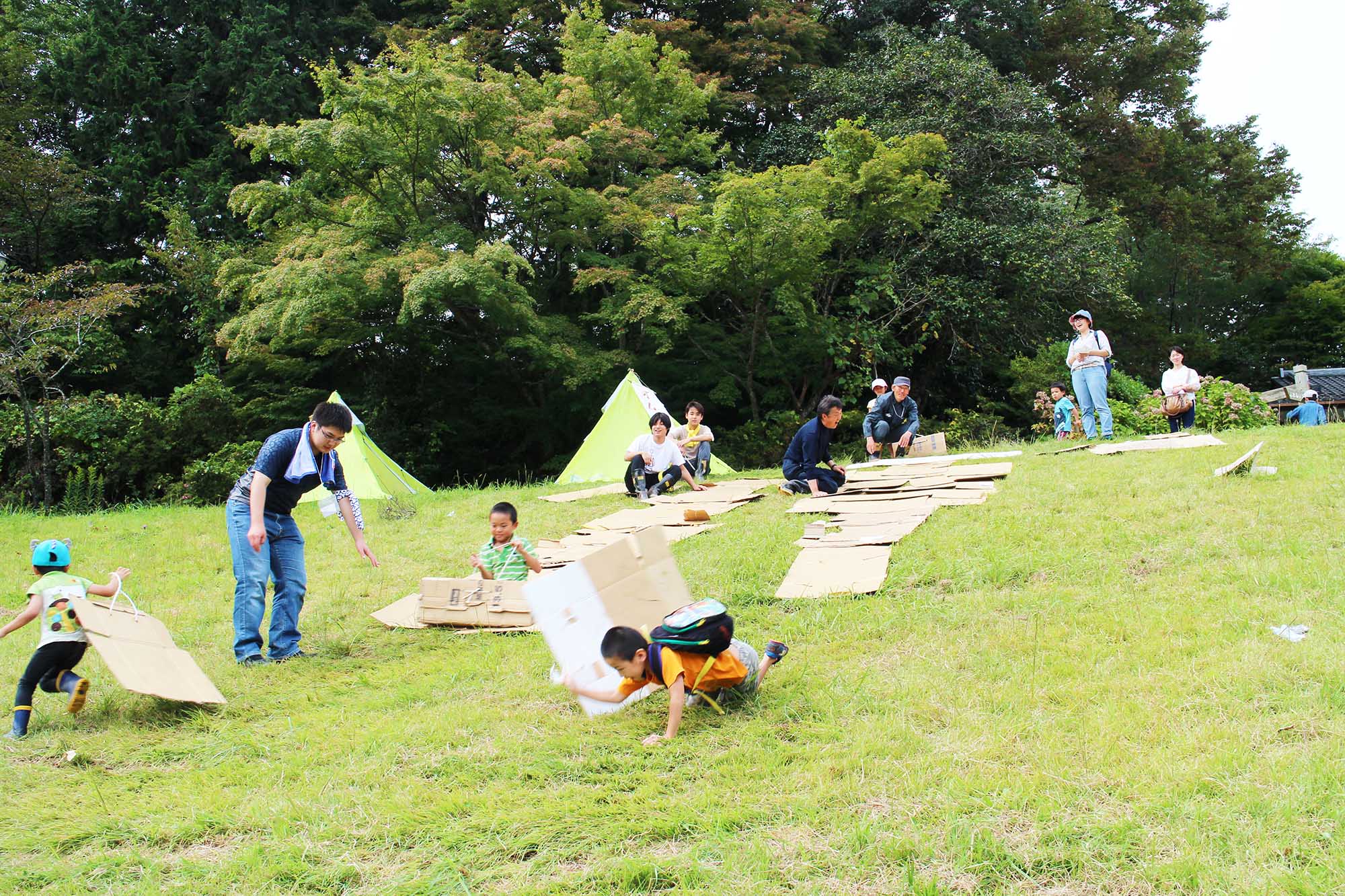 斜面に広げた段ボールの上を子どもたちが勢いよくすべり降り、保護者たちが見守る中で楽しそうに遊んでいる段ボールすべり遊びの写