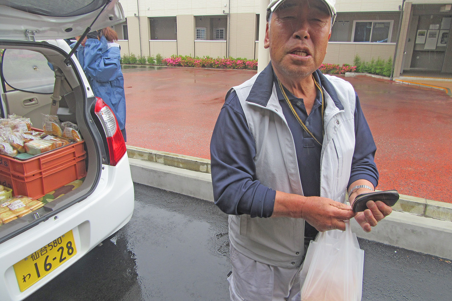 移動販売車の横で中年の男性が購入したパンが入った買い物袋と小銭入れを手に持ってカメラを見ている写真