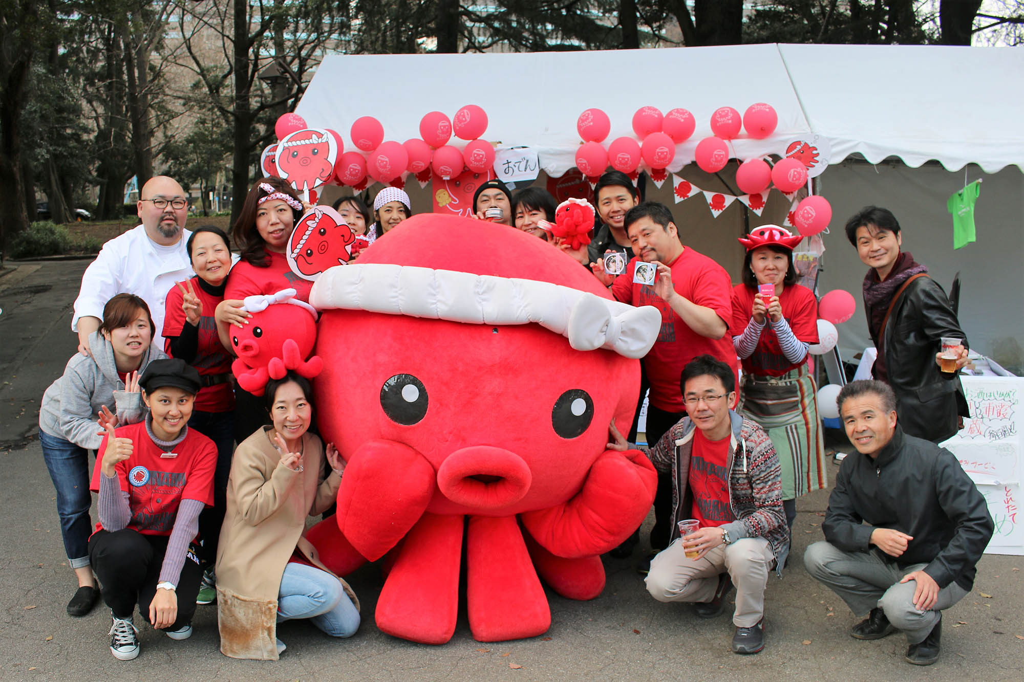 赤いタコのキャラクター「オクトパス君」を囲んで、大勢の大人たちがテントの前で笑顔で集合写真を撮っている写真