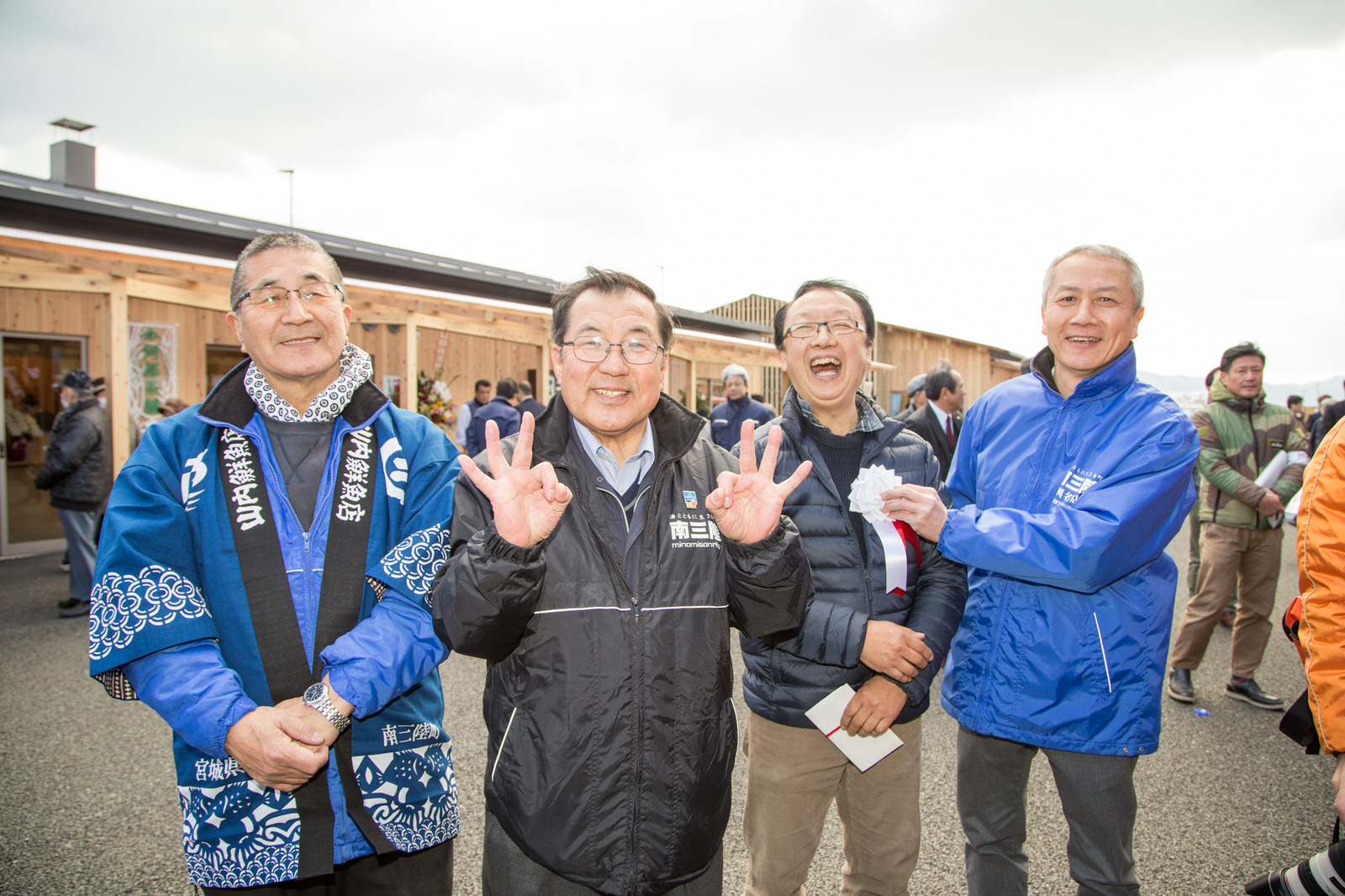 背景には新しく建てられた木造の店舗が見える商店街の屋外で、法被やジャンパーを着た4人の関係者の男性たちが笑顔で並んでいる写真