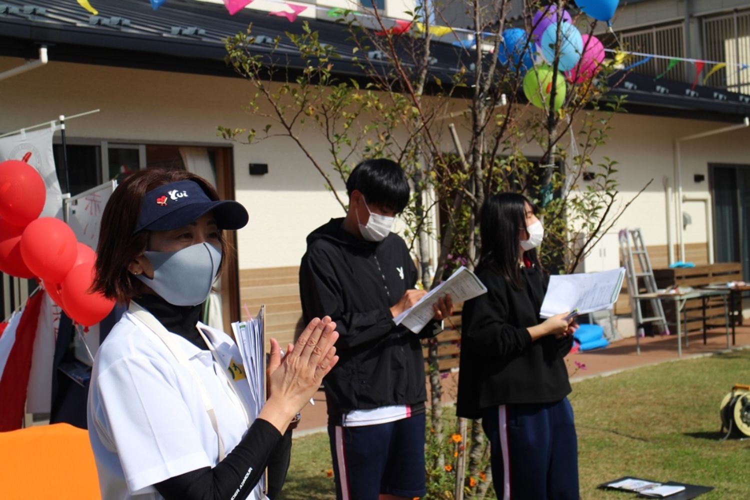 白いポロシャツにサンバイザーとグレーのマスクをつけた女性が手を叩いており、後ろには黒い服を着た男女二人が資料を持って運動会を見ている様子の写真