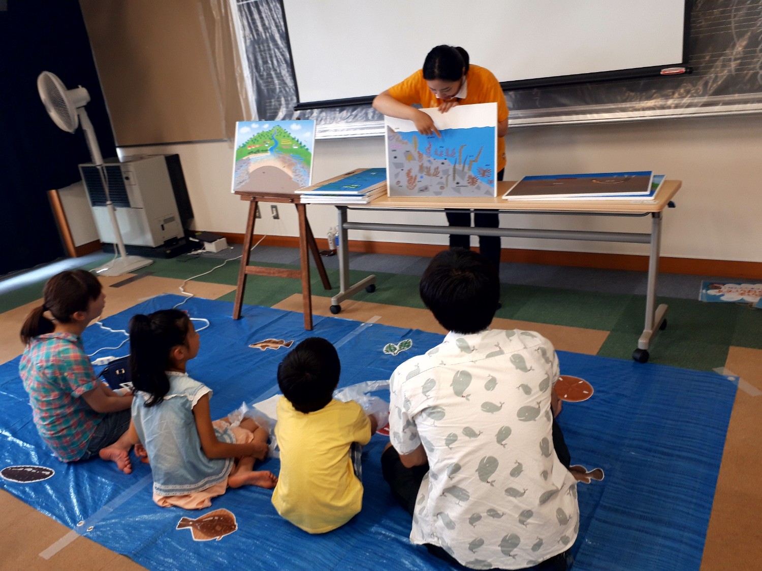 床に敷かれた青いビニールシートの上に子どもたちと大人が座り、前に立つ女性講師が絵を使って説明する海の生き物の話を熱心に聞いている様子の写真