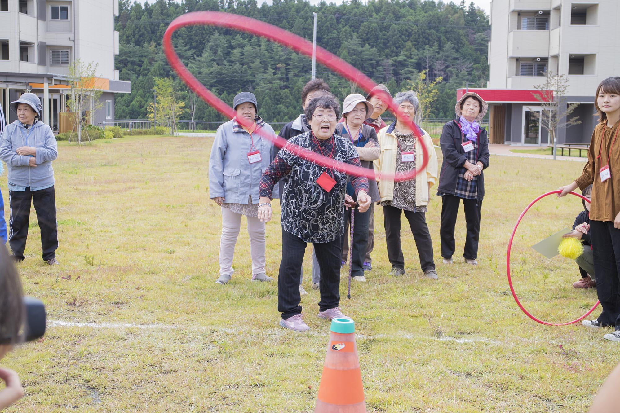 女性が赤い輪投げをオレンジ色のコーンに向かって投げており、周囲にいる他の参加者たちはその様子を笑顔で見守っている、屋外での輪投げ競技中の写真