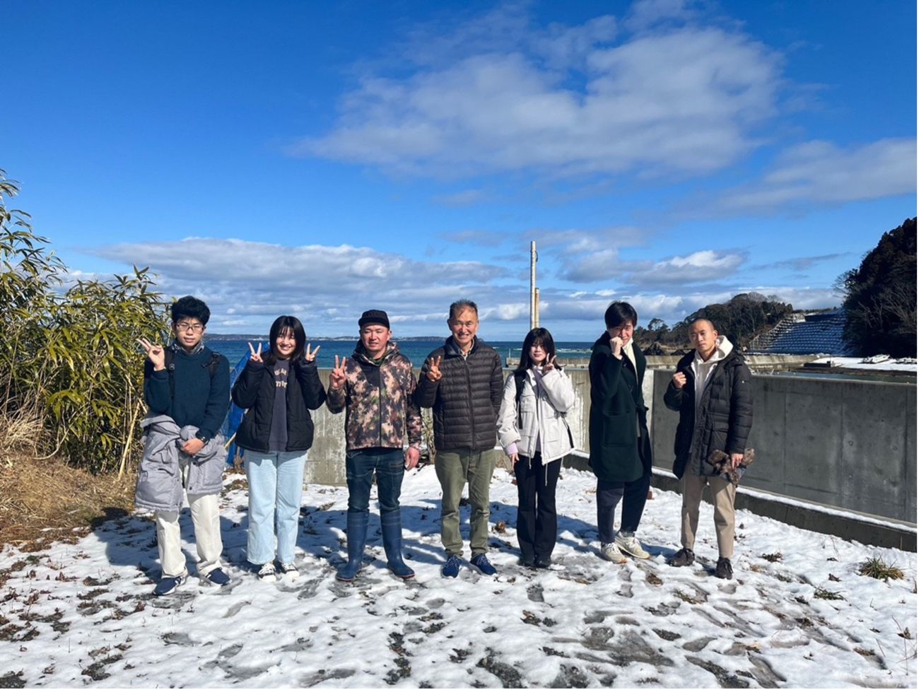 背景には青い空と海が見え、雪が積もった地面の上に7人の男女が並んで立っている屋外の写真