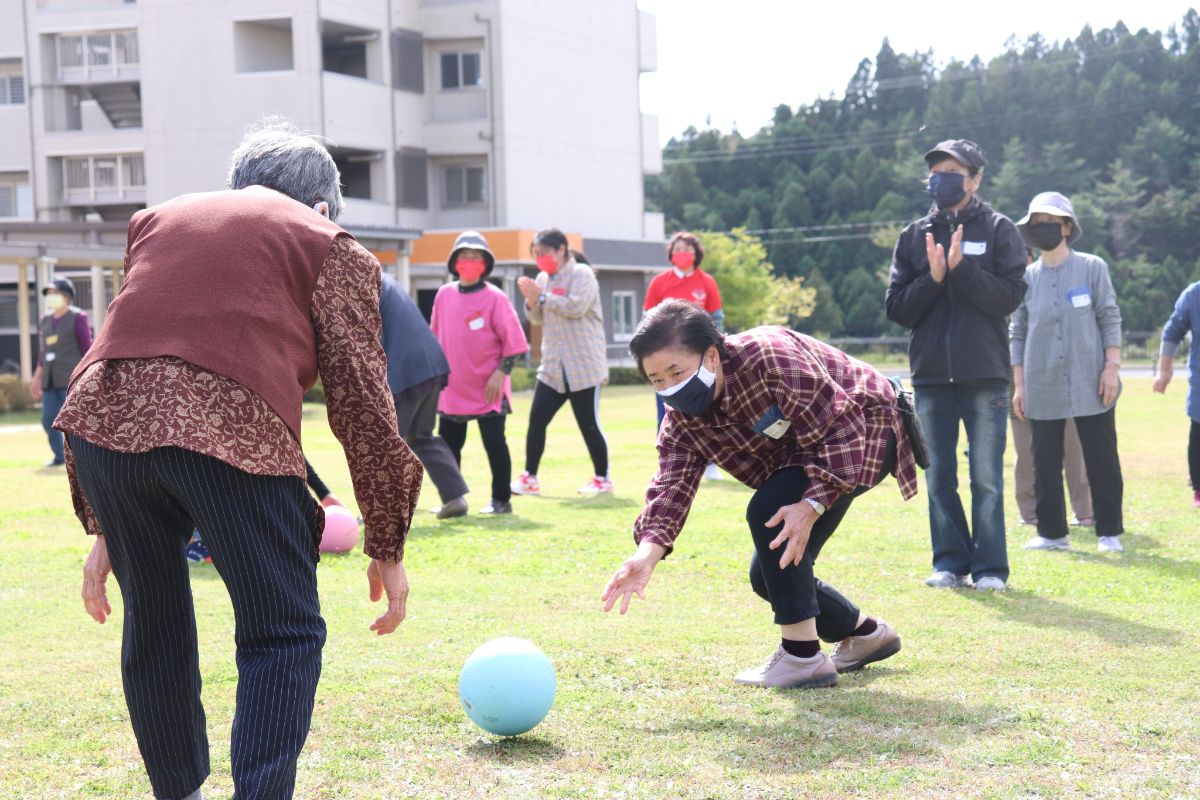 チェックのシャツを着た高齢の女性が芝生の上でボールに手を伸ばし、周りで見守る人々が拍手している「玉転がしリレー」の競技を写した写真