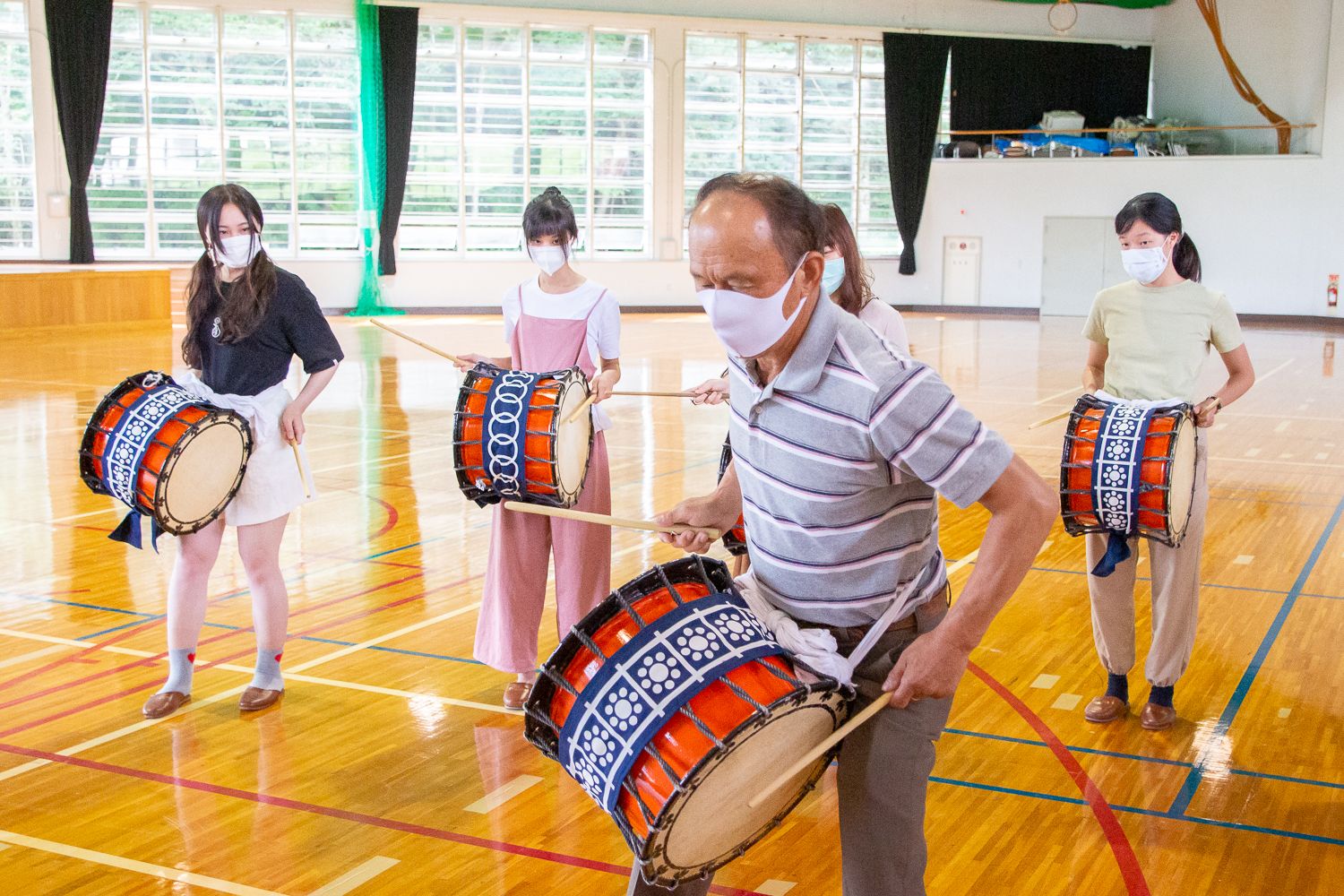 腰に太鼓をつけ、両手にバチを持って立っている4人の参加者たちの前で、太鼓を腰に付けた村岡会長が太鼓のたたき方の見本を見せている様子の写真
