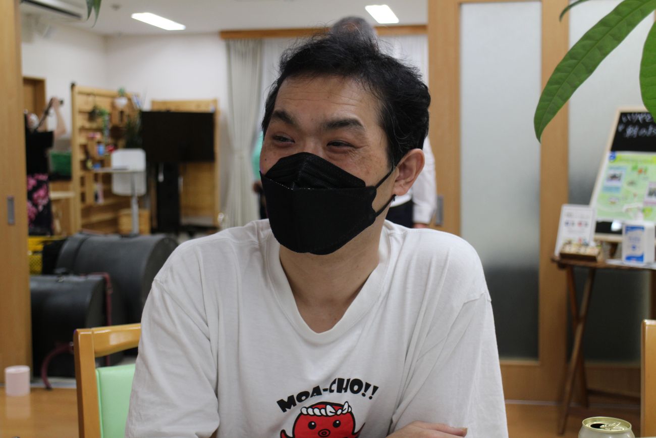 黒いマスクを着用し白いTシャツを着ている男性が室内のテーブルに座り笑っている様子の写真