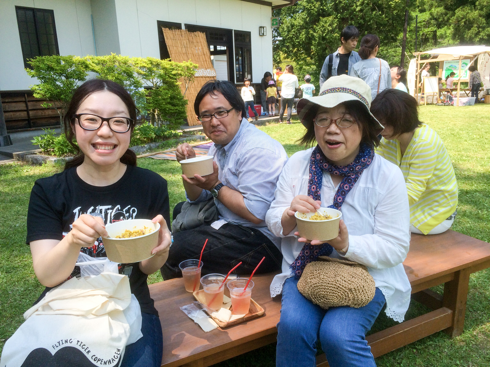 人々でにぎわう芝生広場のベンチで、テイクアウト風の容器に入った食事を手にした3人の男女が笑顔で食事を楽しんでいる写真