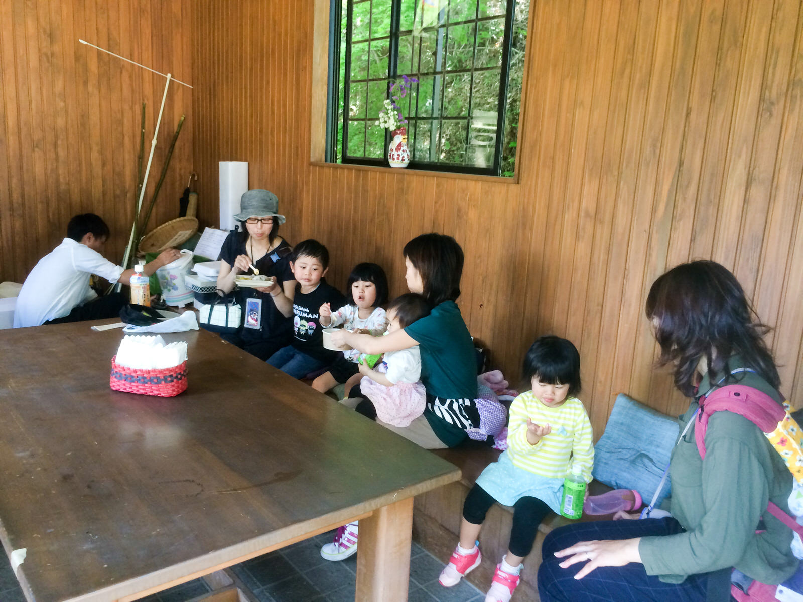 木の壁に囲まれた休憩所のような室内で、大人と子どもが一緒に座ってお弁当を食べてくつろいでいる様子の写真