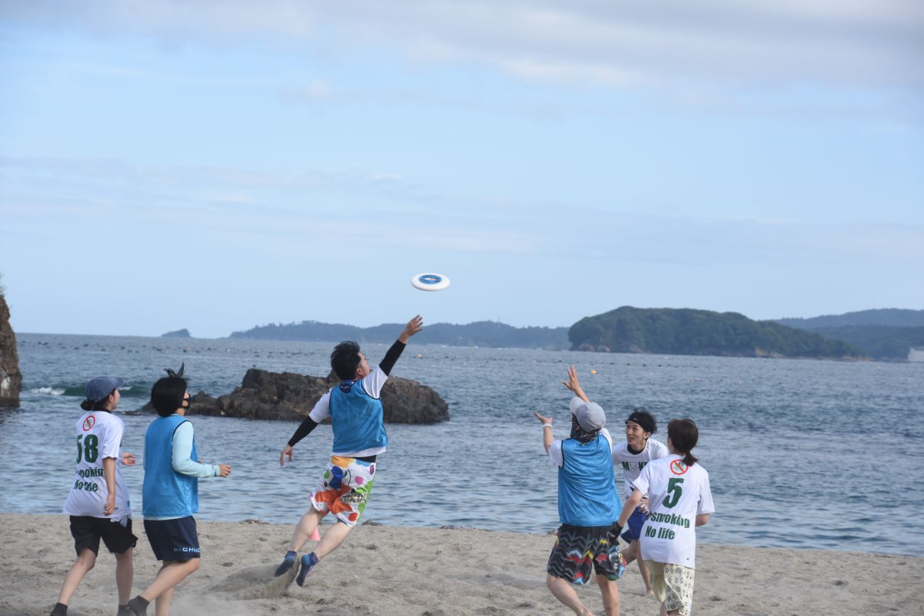 数人の若者たちが砂浜でフリスビーを追いかけ、ジャンプしてキャッチを狙っている様子の写真