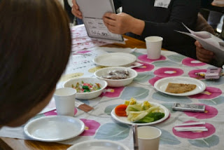 机の上に、漬け物や菓子類が並べられ、参加者が資料を見ながら話し合っている様子の写真