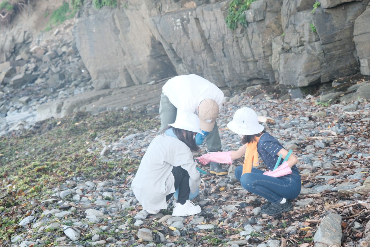 数人の子どもたちが浜辺でしゃがみ、何かを拾って観察しながら清掃活動を行っている様子の写真