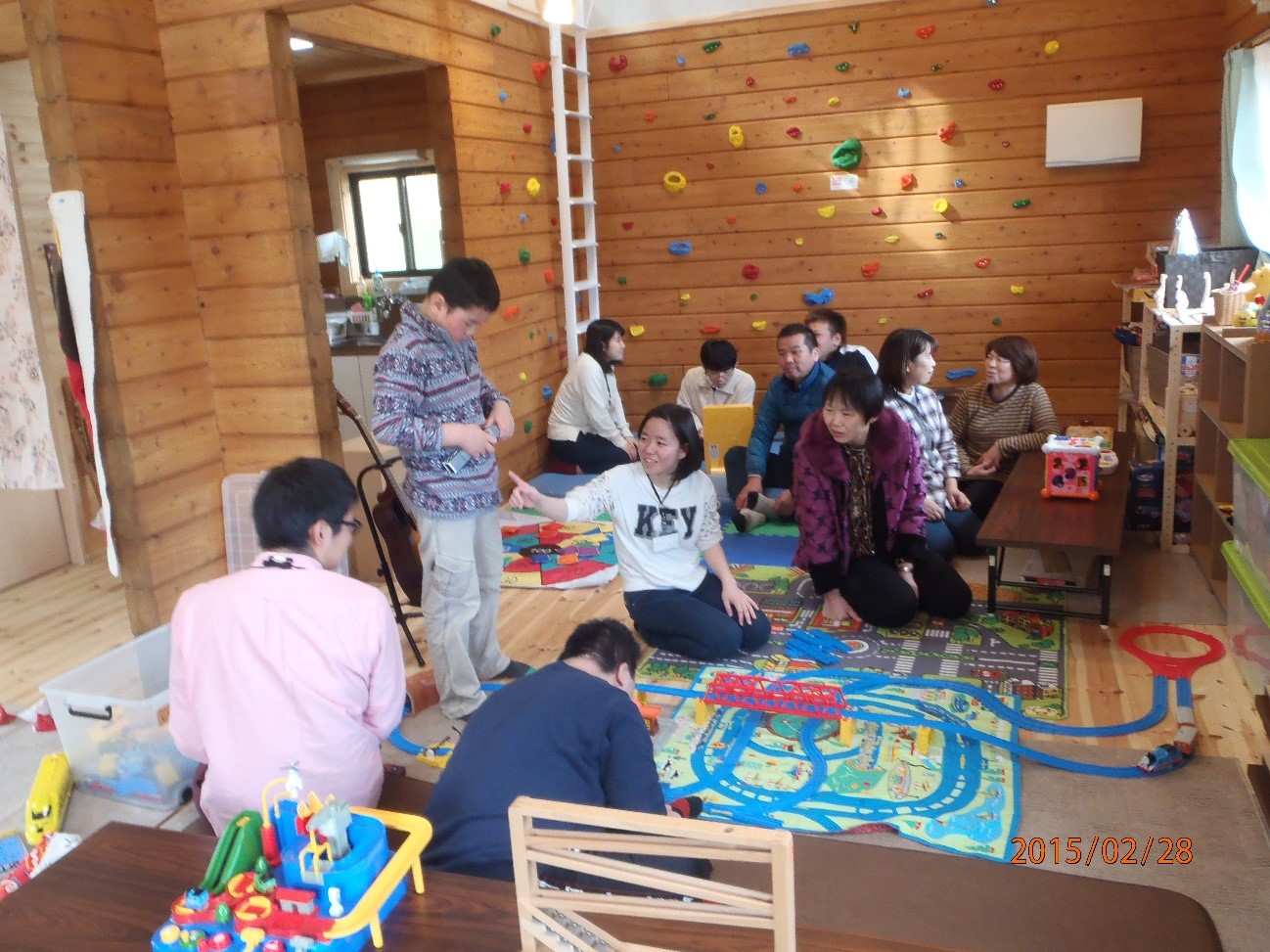 壁の一部がクライミングウォールになっている木造の部屋の中で、床に敷かれたカラフルなプレイマットの上で子供たちと大人たちが一緒に遊んでいる様子が写っている写真