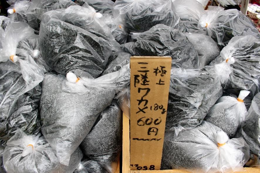 段ボールを細長く切った札に値段と内容量が書かれ、黒いワカメが詰まった多数のビニール袋が無造作に積まれているワカメ販売の写真